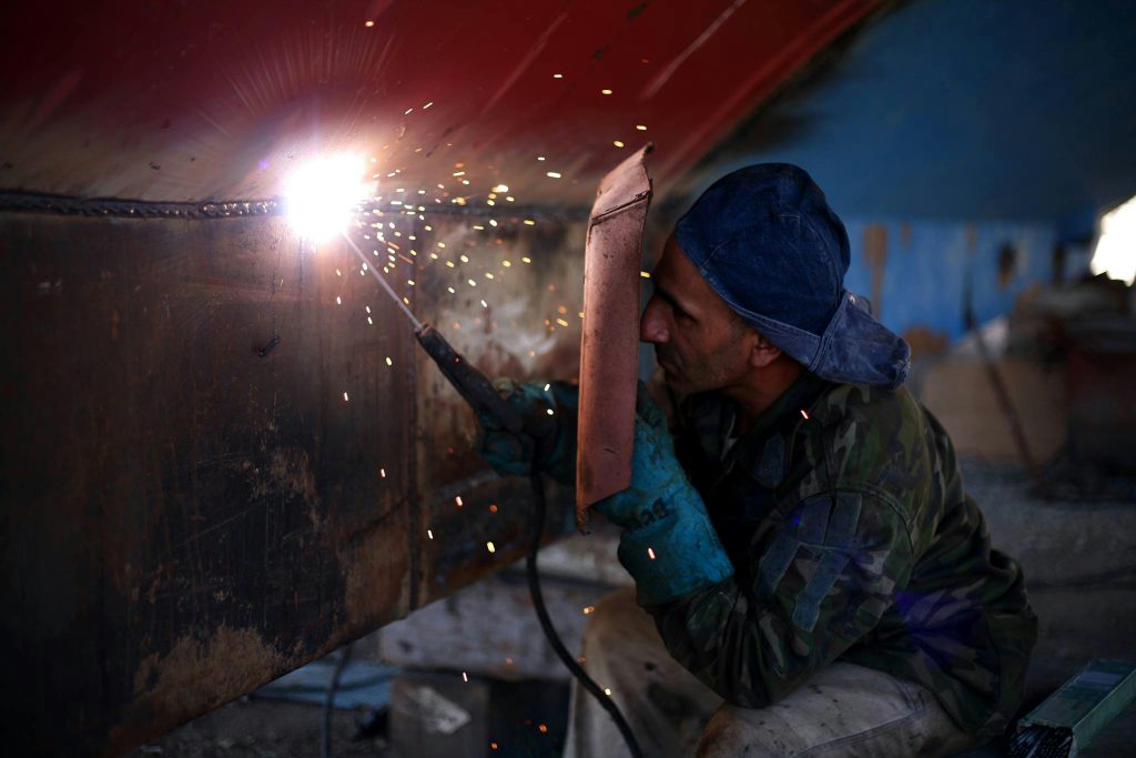 A welder in protective gear works indoors, generating sparks with a welding torch.